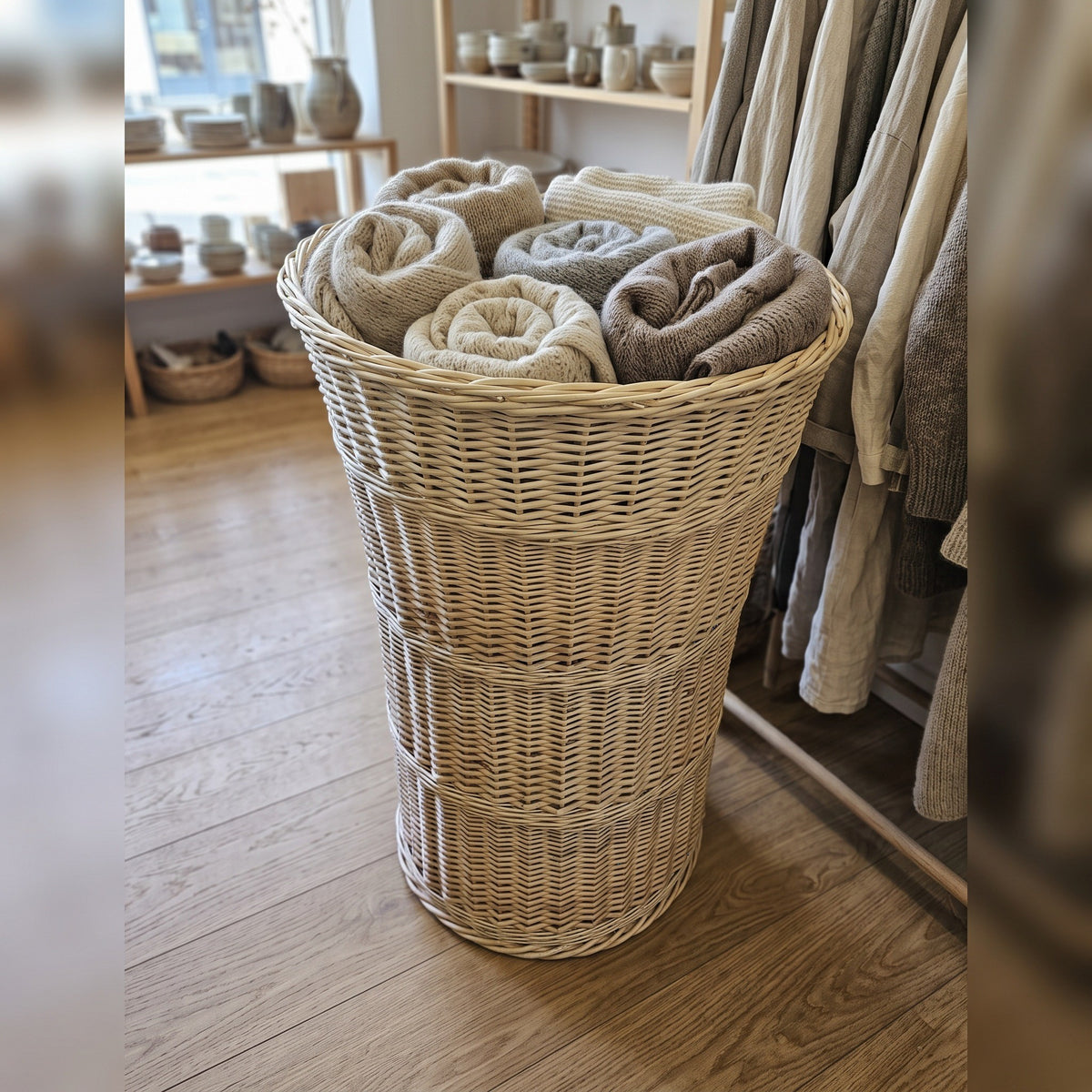 Tall tapered willow basket holding rolled wool blankets in a home goods shop beside a rail of linen clothing