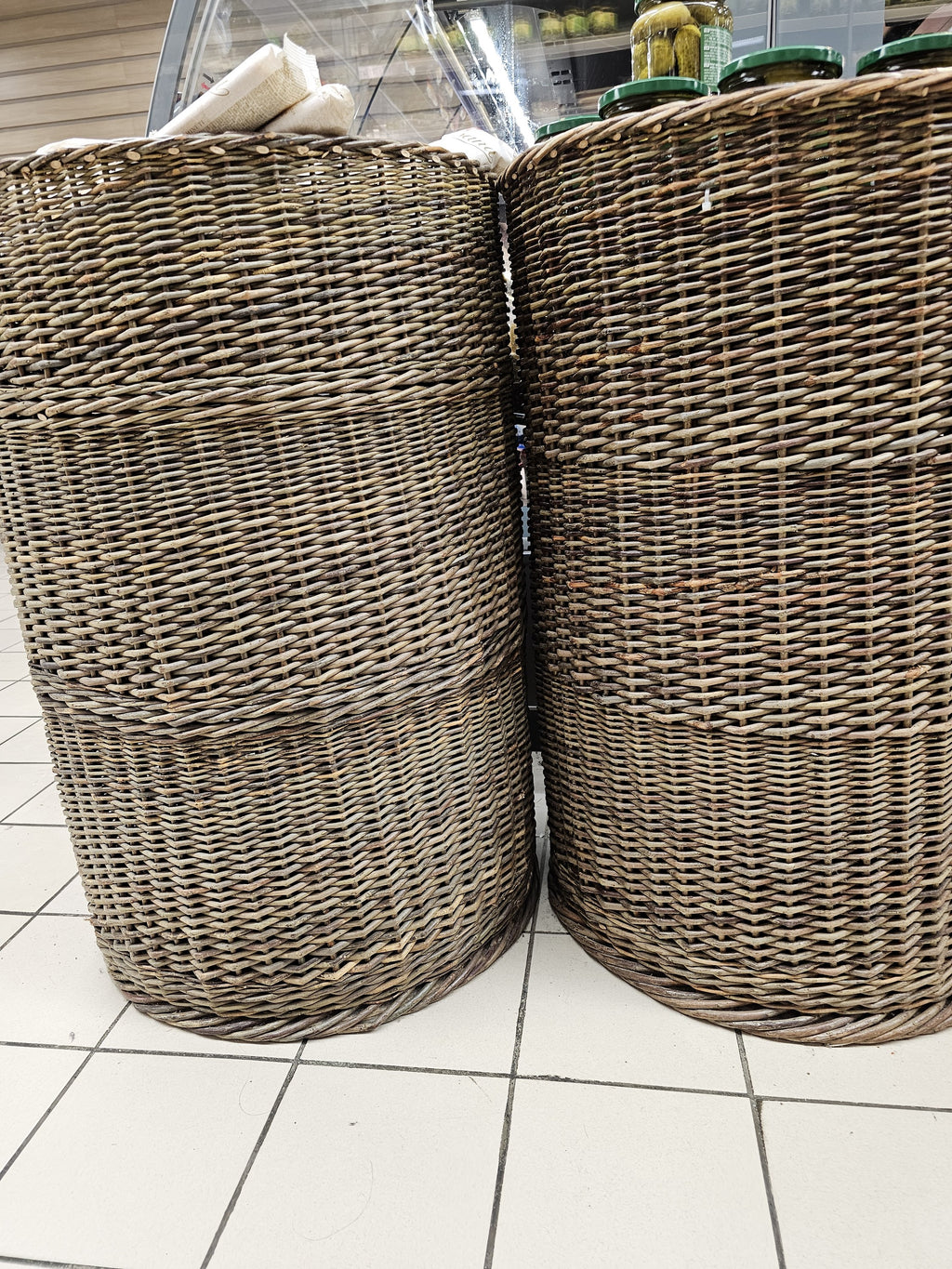 Two tall handwoven willow display baskets standing side by side on a white tiled shop floor