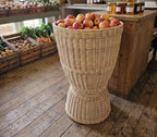 Tall hourglass willow display basket filled with apples on the wooden floor of a farm shop beside crates of vegetables