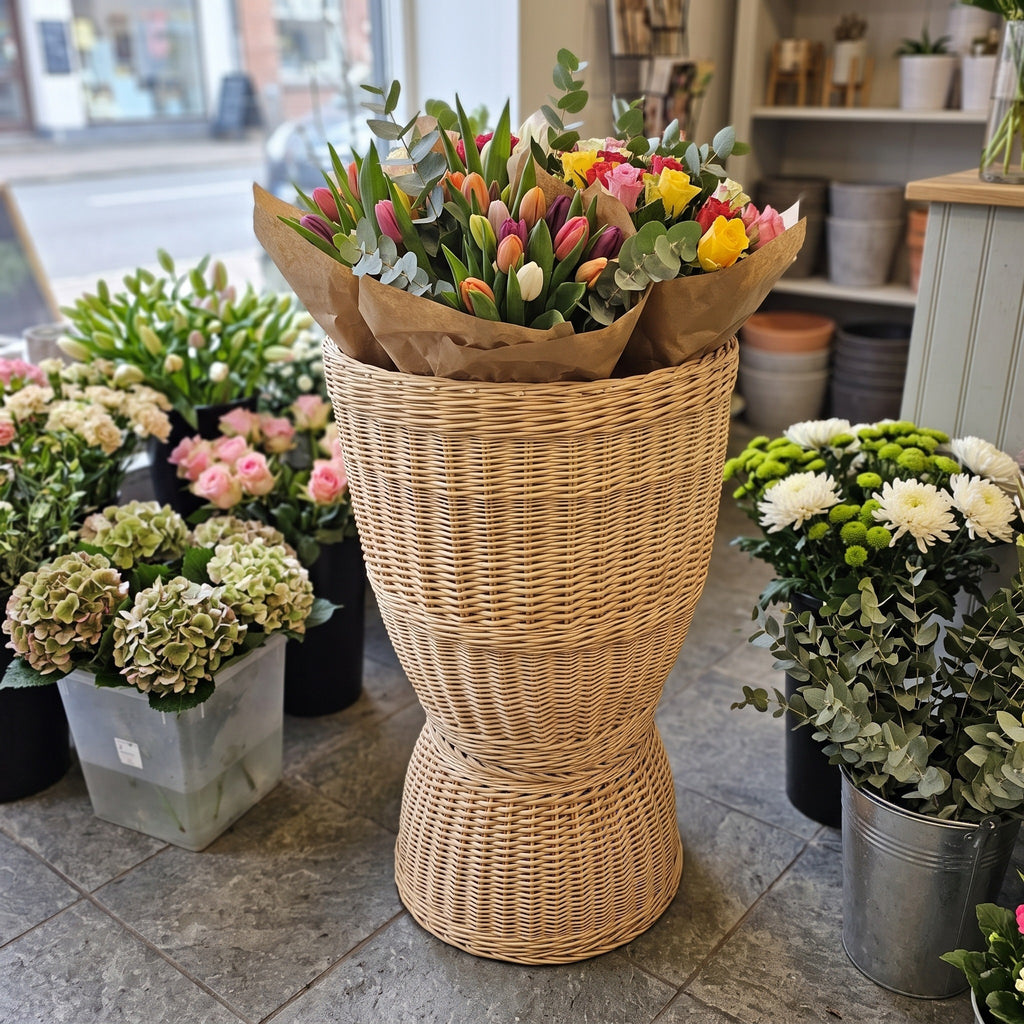 Tall wicker floor display basket holding wrapped tulip bouquets among potted plants and flower buckets in a florist shop