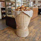 Hourglass wicker display stand lined with linen and piled with cinnamon buns and croissants in front of a bakery counter