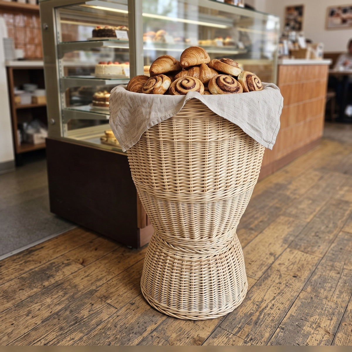 Hourglass wicker display stand lined with linen and piled with cinnamon buns and croissants in front of a bakery counter