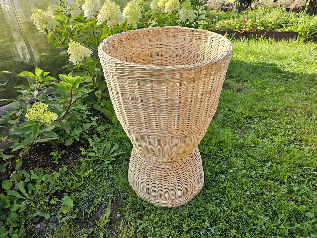 Willow pedestal display basket with an hourglass shape, flared top and wide foot, standing on the grass in a garden