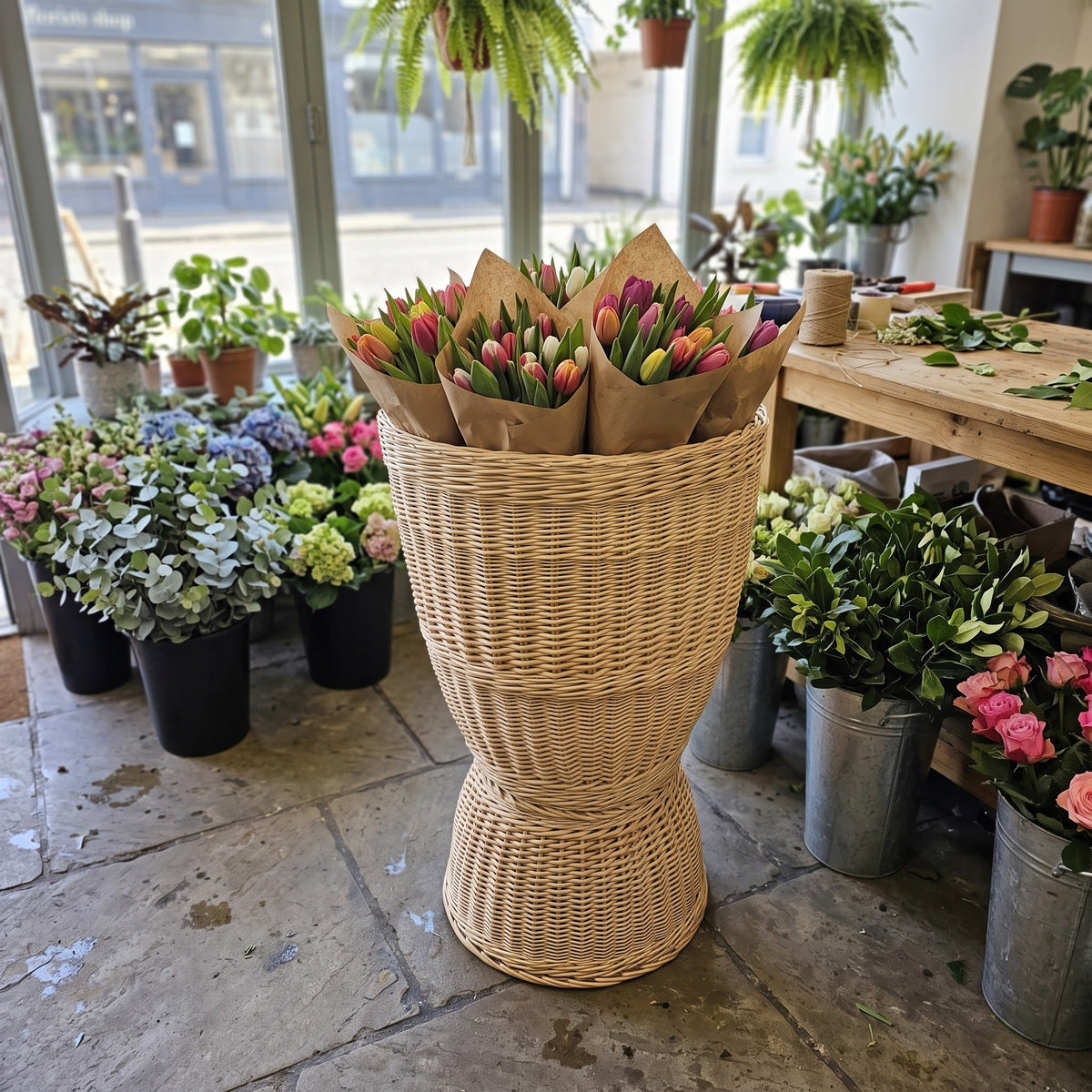 Willow floor display basket holding paper-wrapped tulip bunches in a bright flower shop with hydrangeas and potted plants