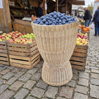 Tall hourglass wicker basket heaped with plums at an outdoor farmers market next to crates of apples on cobblestones