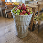 Tall tapered willow floor basket filled with red and green apples on a wooden floor in a farm shop