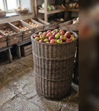 Tall round willow display basket packed with fresh apples on a stone floor in a farm shop with produce crates