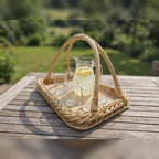 Handwoven willow serving tray with handles carrying a glass jug of lemonade and drinking glasses on a wooden garden table