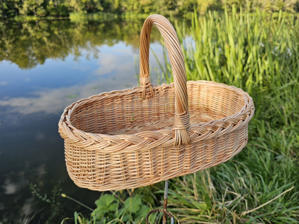 Low oval wicker trug with a tall handle and braided rim, standing on the grassy bank of a calm pond on a summer evening
