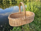 Low oval wicker trug with a tall handle and braided rim, standing on the grassy bank of a calm pond on a summer evening