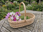 Small willow flower basket with handle holding cosmos and daisies on a wooden garden table in a flowering cottage garden