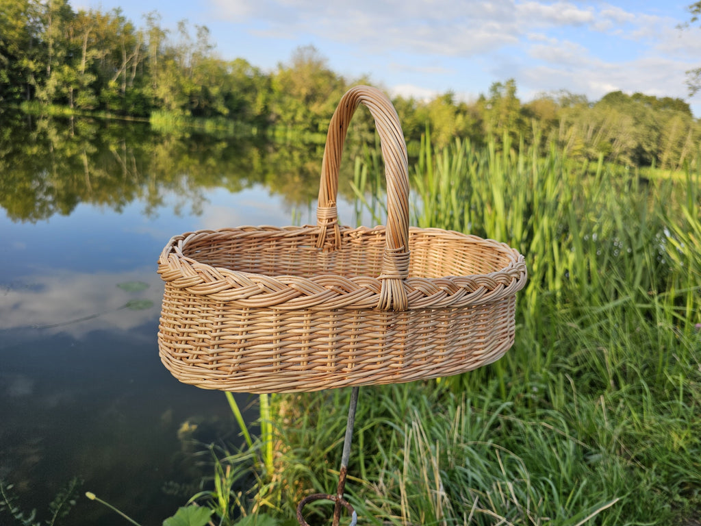 Small wicker gathering trug with a braided rim and tall handle on the grass by a pond reflecting the evening sky
