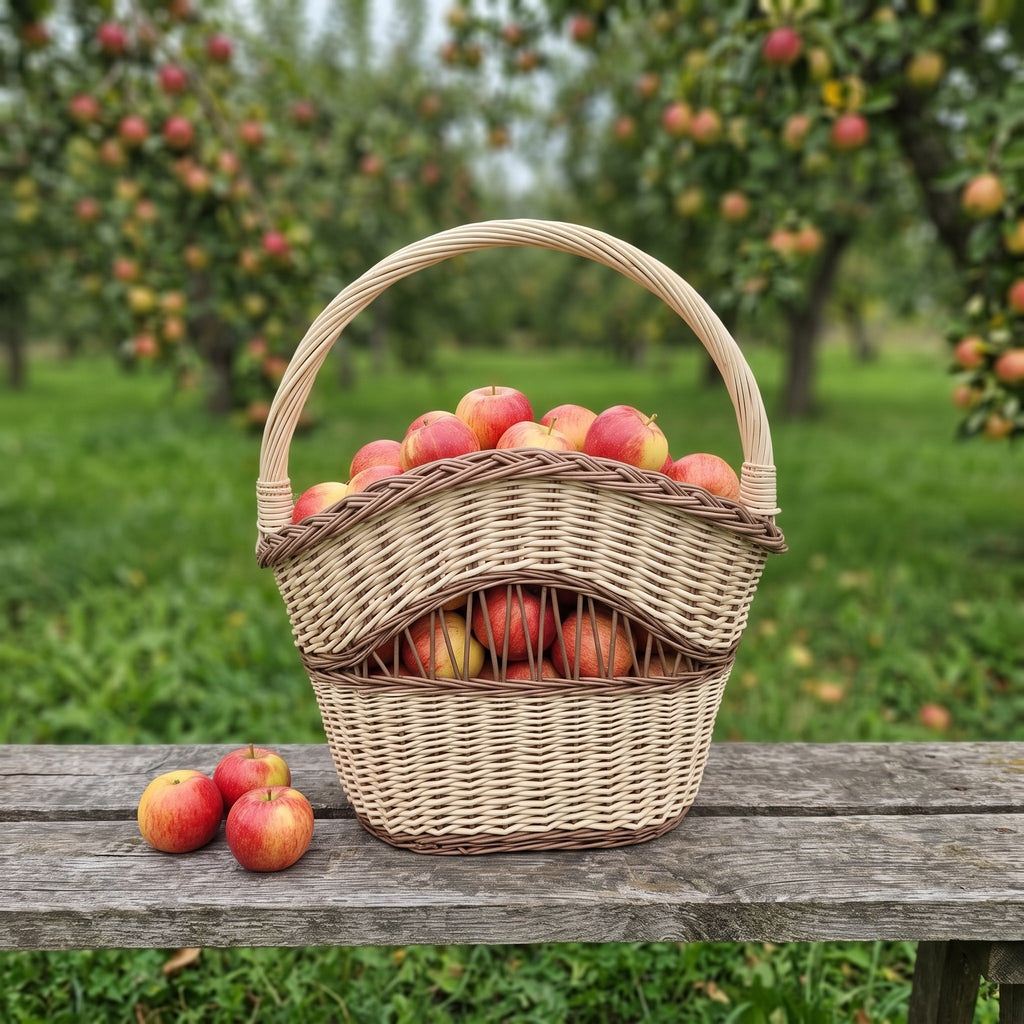 Handwoven willow apple basket with an arched handle and open-work front, filled with fresh apples on a bench in an orchard