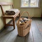 Tall handwoven wicker basket used as a knitting basket with balls of yarn and needles, next to a wooden bench by the window