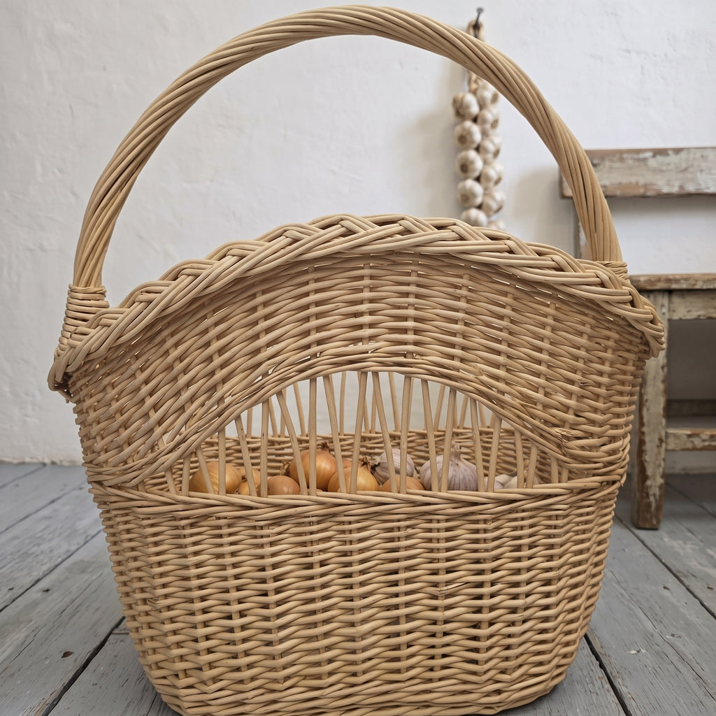 Tall wicker basket with handle and braided rim, filled with onions behind the open-work band, beside a rustic wooden stool