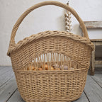 Tall wicker basket with handle and braided rim, filled with onions behind the open-work band, beside a rustic wooden stool
