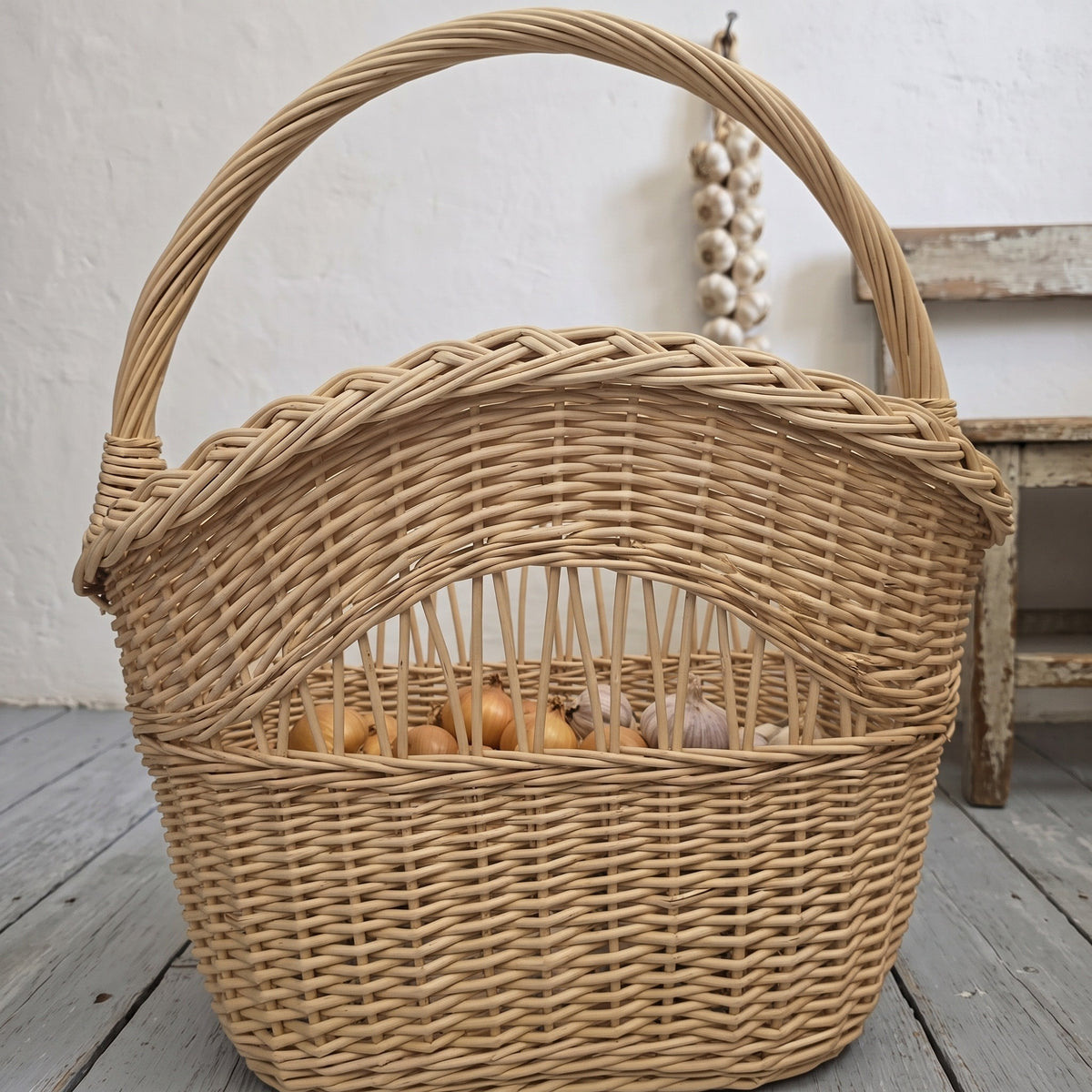 Tall wicker basket with handle and braided rim, filled with onions behind the open-work band, beside a rustic wooden stool