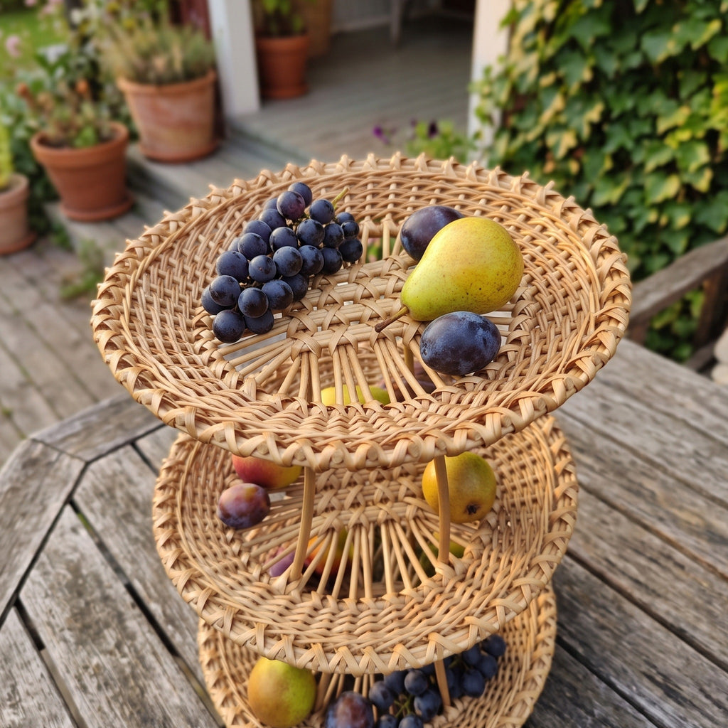 Tiered wicker fruit basket stand filled with grapes, plums and pears on a wooden patio table among potted plants