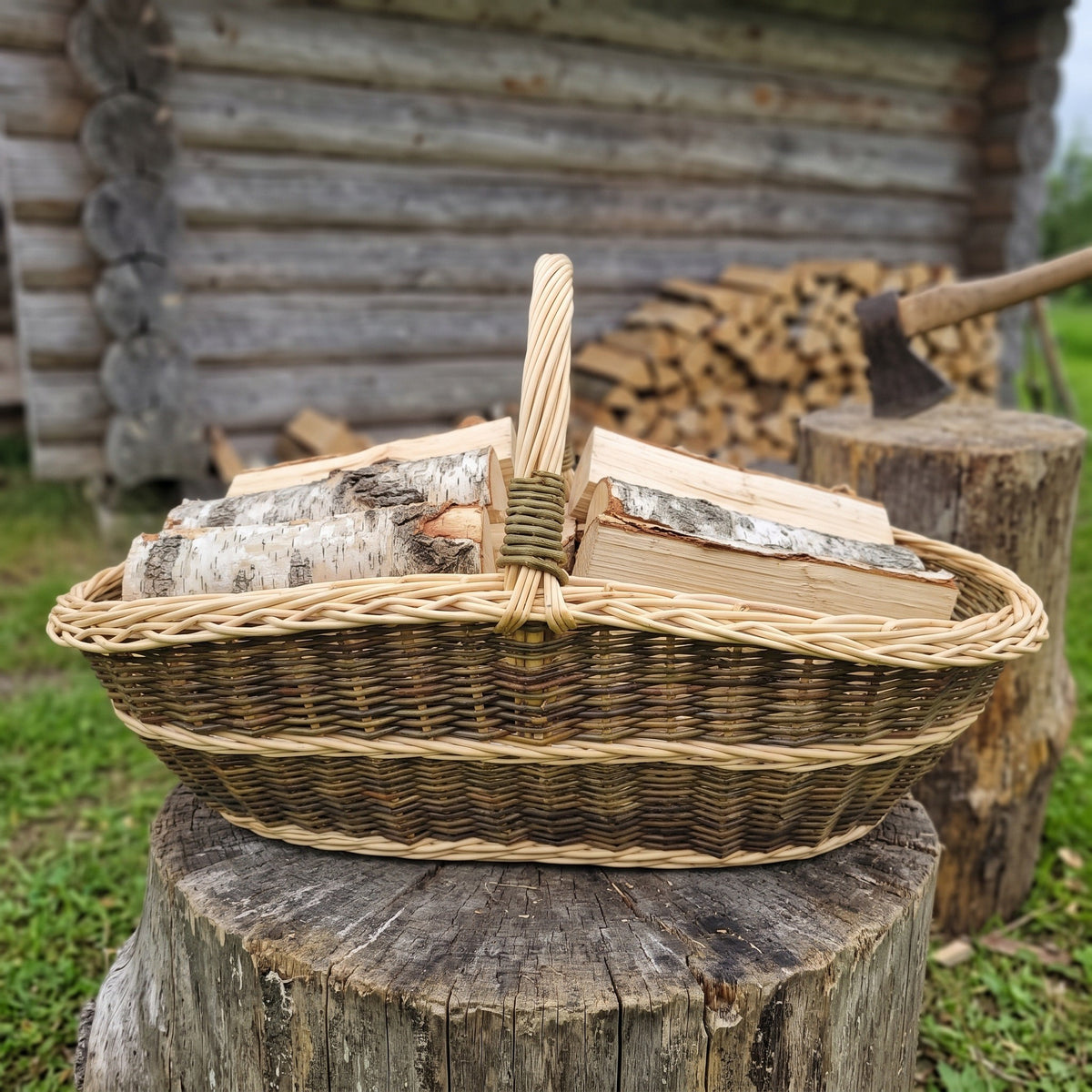 Extra large handwoven willow firewood basket with a single arched handle, filled with split birch logs and standing on a tree stump beside a woodpile.