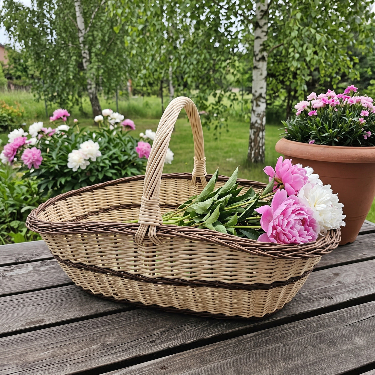 Extra large willow trug basket with a twisted handle holding fresh peonies on a garden table in a summer garden with birch trees