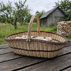 Extra large wicker log carrier trug with handle holding birch logs on a table in an orchard beside a stack of firewood