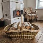 Handwoven willow log basket filled with split firewood, standing on a wooden floor in front of a tiled stove with a crackling fire.