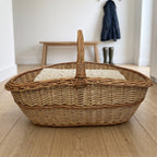 Extra large wicker basket with a tall handle holding a folded knitted blanket on a wooden hallway floor, a bench, coat and boots behind.