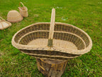 Handwoven willow basket seen from above on a tree stump in a garden, showing the deep oval interior and the bound arched handle, with other baskets behind.