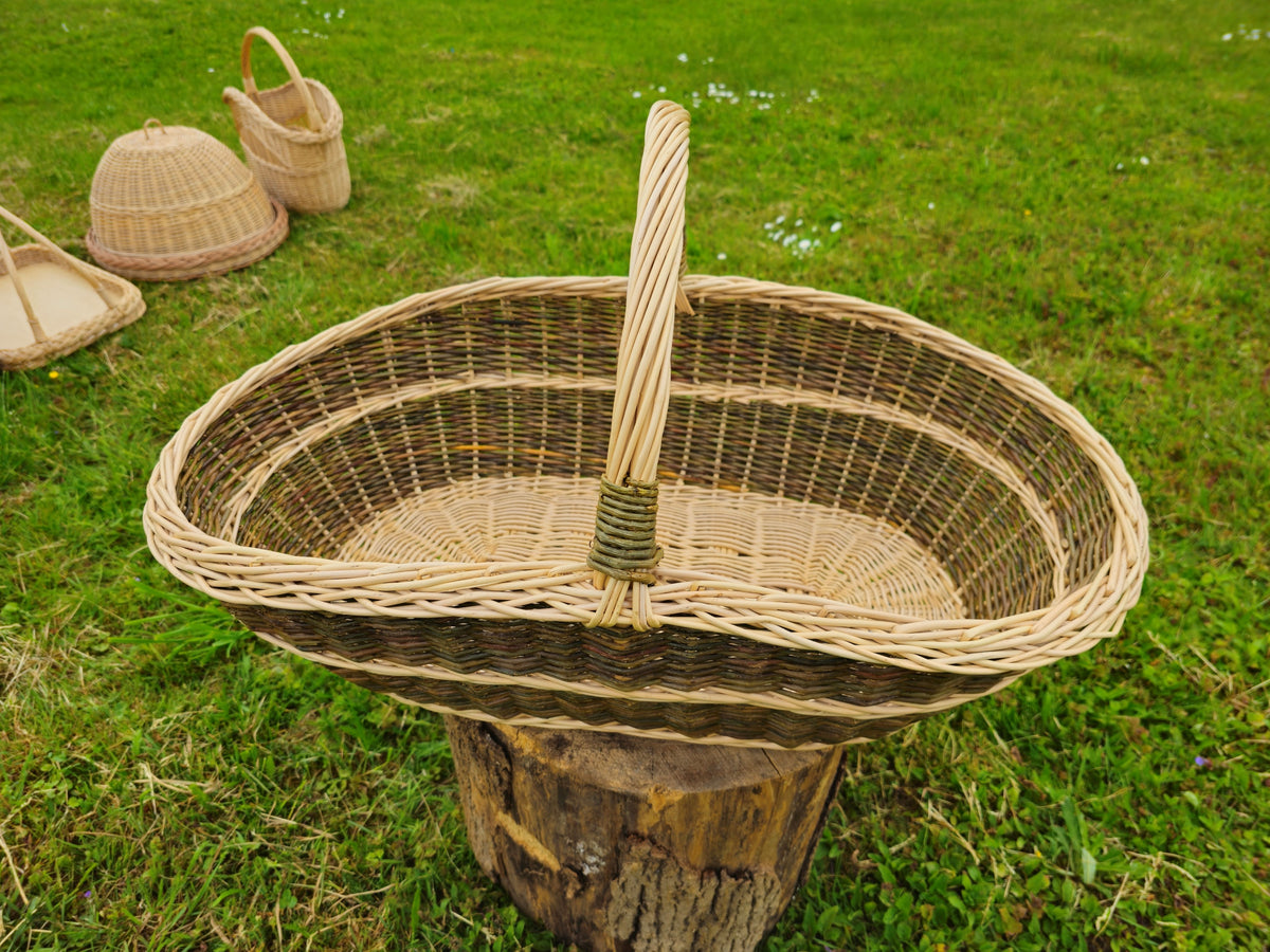 Handwoven willow basket seen from above on a tree stump in a garden, showing the deep oval interior and the bound arched handle, with other baskets behind.