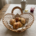 Round wicker fruit bowl with two looped handles used as a bread basket for rolls on a linen cloth at a breakfast table