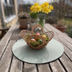 Small handwoven willow Easter basket bowl with handles holding dyed eggs on moss, on a garden table with daffodils