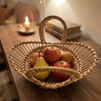 Wicker fruit bowl with handles filled with apples and a pear on a wooden sideboard beside books and a lit candle