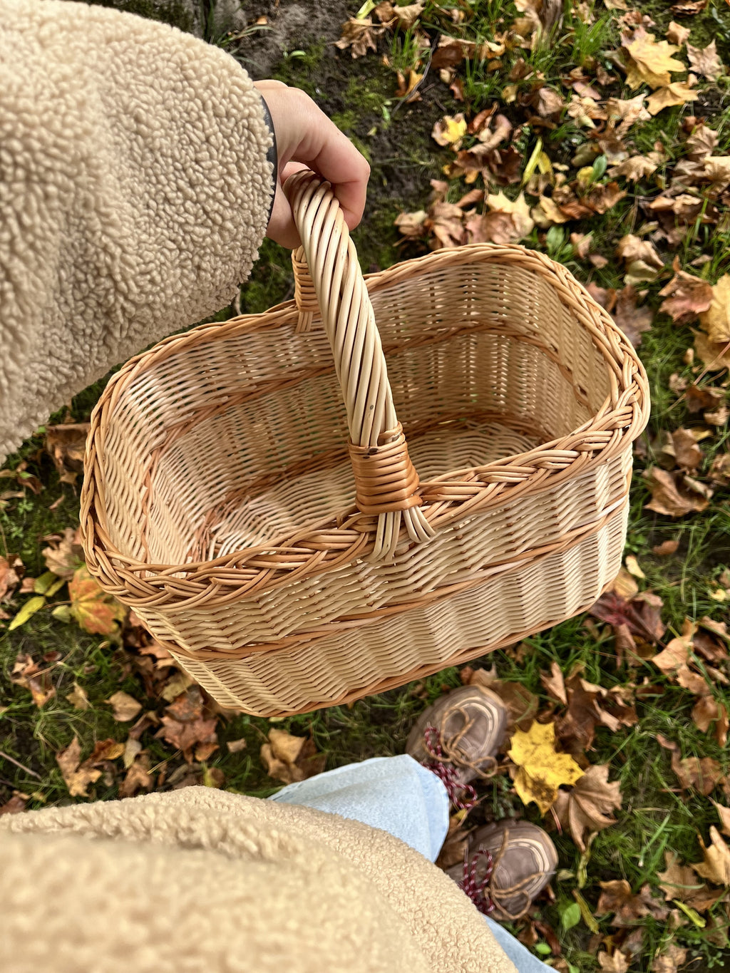 Hand holding the empty willow basket by the handle over autumn leaves and boots, seen from above