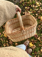 Hand holding the empty willow basket by the handle over autumn leaves and boots, seen from above