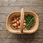 Top view of a wicker basket with handle holding fresh eggs on straw and a bunch of rosemary on a wooden table
