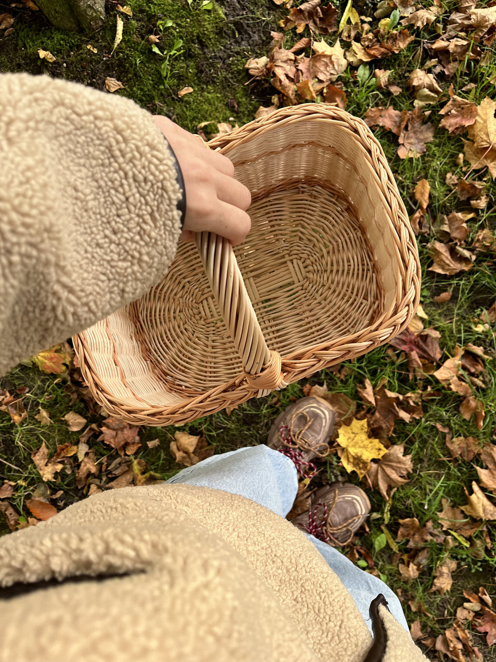 Hand holding an empty rectangular wicker basket by its handle above fallen autumn leaves, showing the woven base