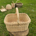Empty rectangular wicker basket with braided rim and tall handle on a tree stump, with more woven baskets on the grass