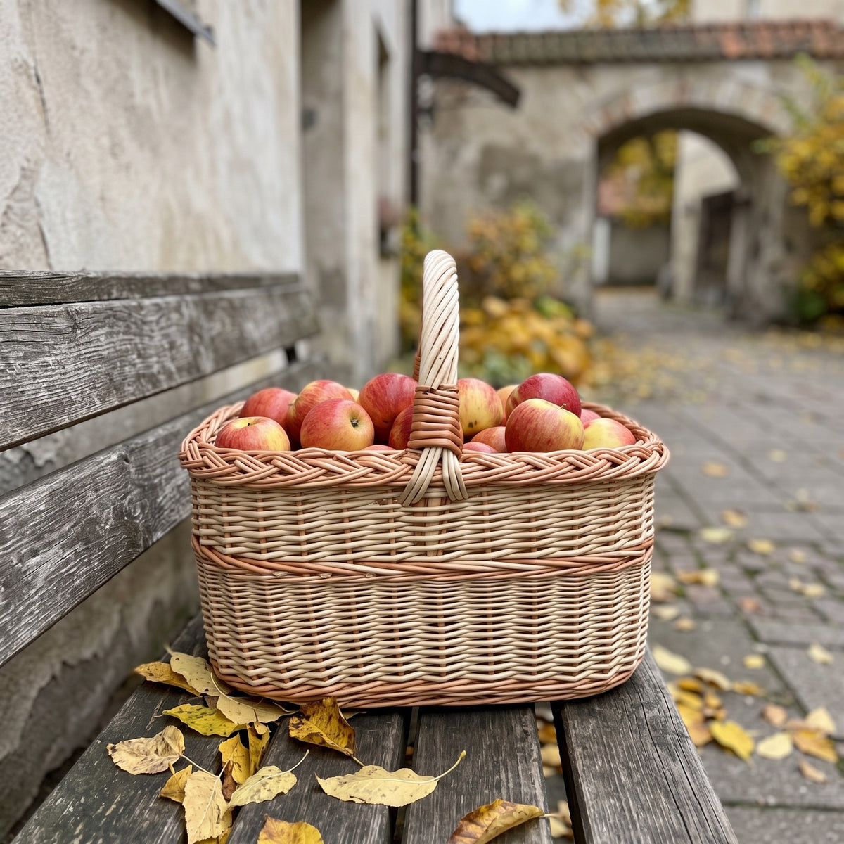 Wicker basket with handle full of apples on an old wooden bench in a cobbled courtyard with autumn leaves