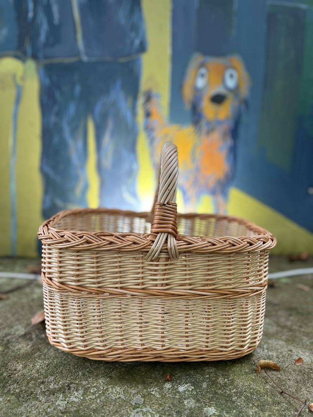 Rectangular willow basket with braided rim and handle on a stone ledge in front of a painted dog mural