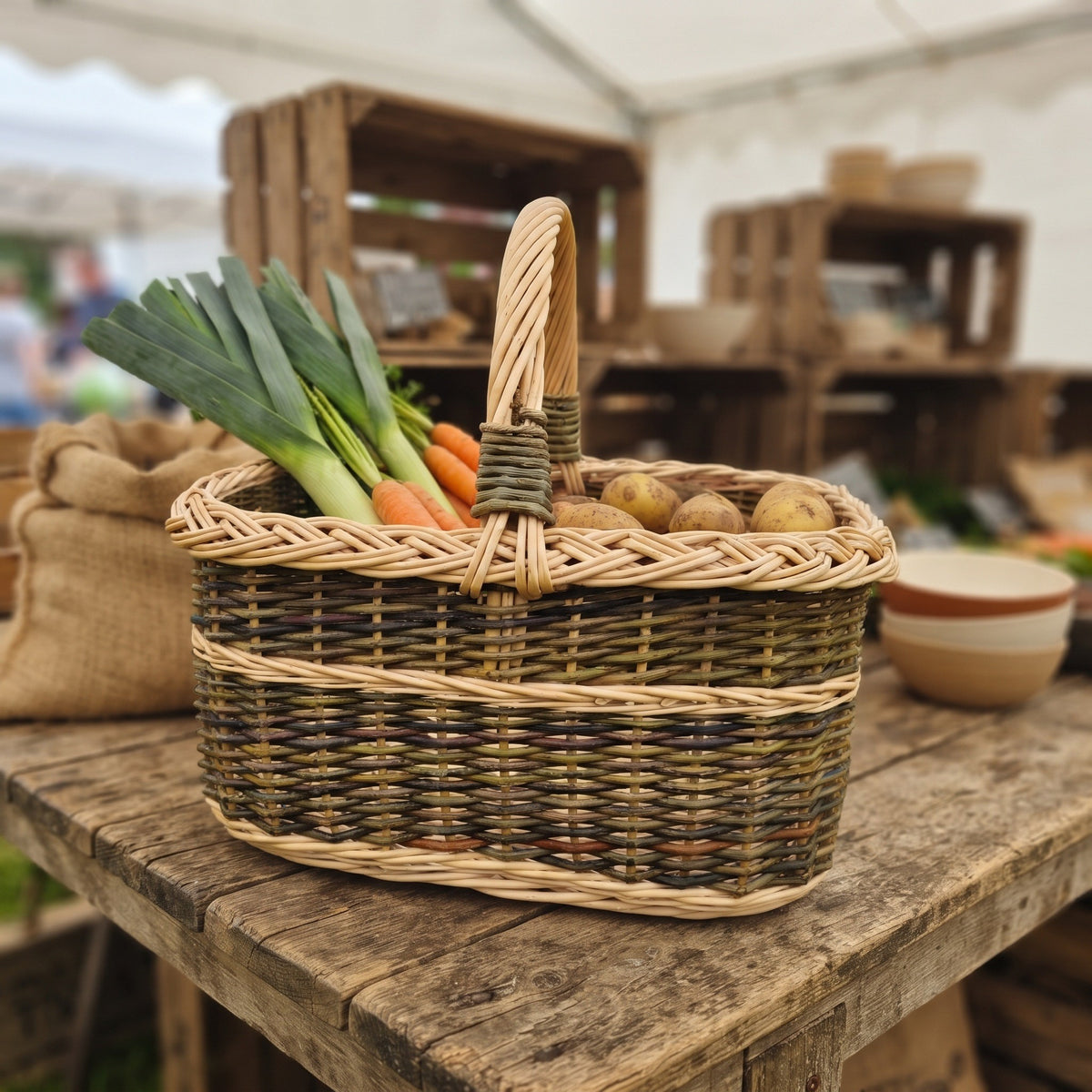 Wicker market basket with handle carrying leeks, carrots and potatoes on a rustic wooden table at a farmers market stall