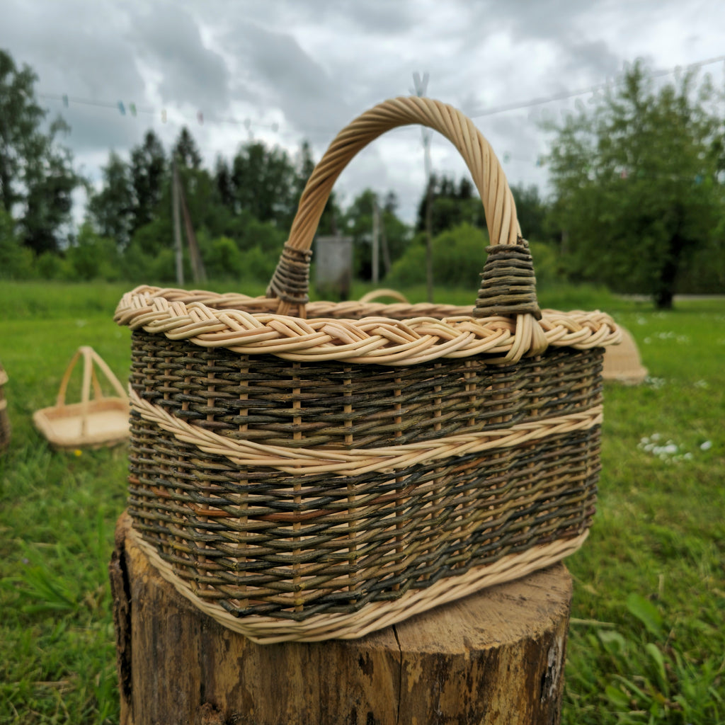 Front view of a handwoven willow carrier basket with a high arched handle on a wooden stump under a cloudy sky