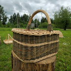 Front view of a handwoven willow carrier basket with a high arched handle on a wooden stump under a cloudy sky