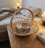 Small wicker basket with handle used as a bedside catch-all, holding a candle, a ring dish and reading glasses on a wooden nightstand
