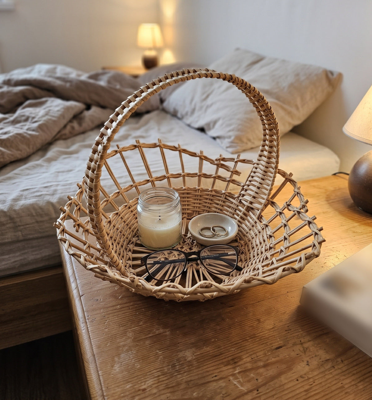 Small wicker basket with handle used as a bedside catch-all, holding a candle, a ring dish and reading glasses on a wooden nightstand