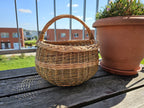 Round wicker gathering basket with a single arched handle and a woven band around the body, on a balcony table next to a flowerpot