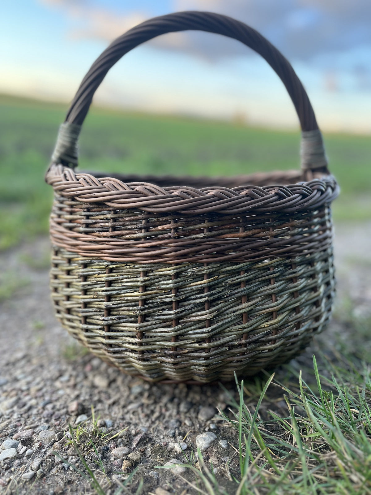 Round willow basket with braided rim and arched handle on a gravel path beside a green field