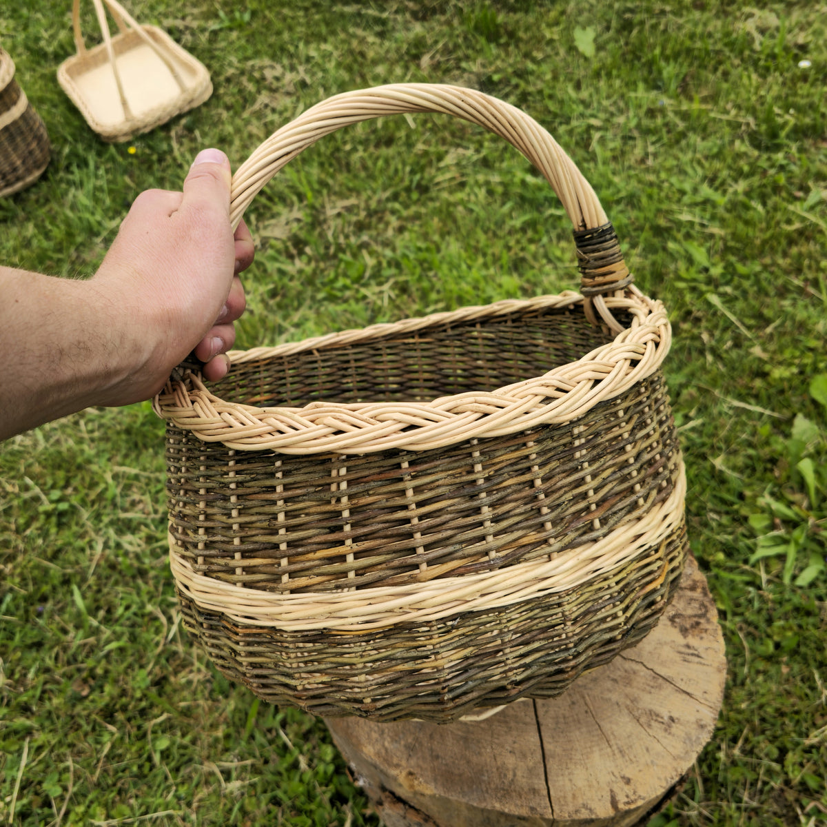 Round foraging basket handwoven from willow held by its arched handle above a tree stump on the grass, finished with a braided rim