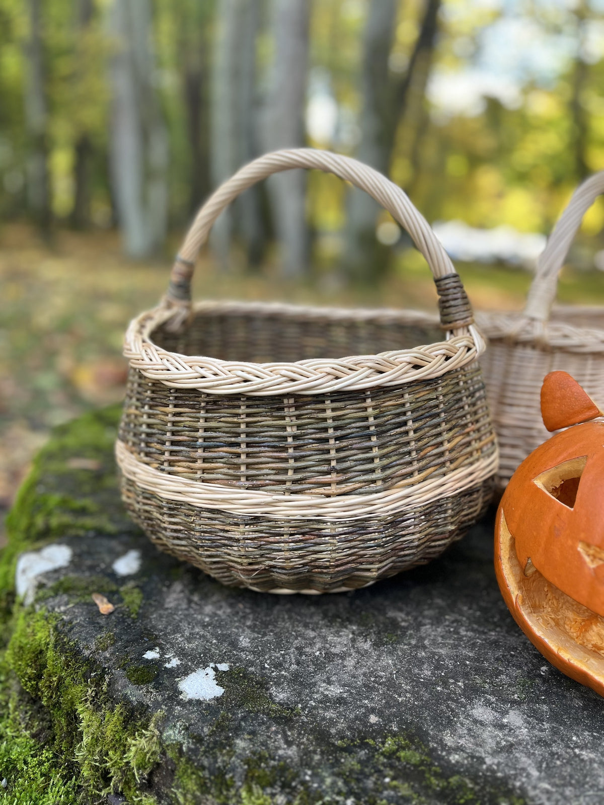 Round wicker basket with braided rim and handle on a mossy stone next to a carved pumpkin and another basket