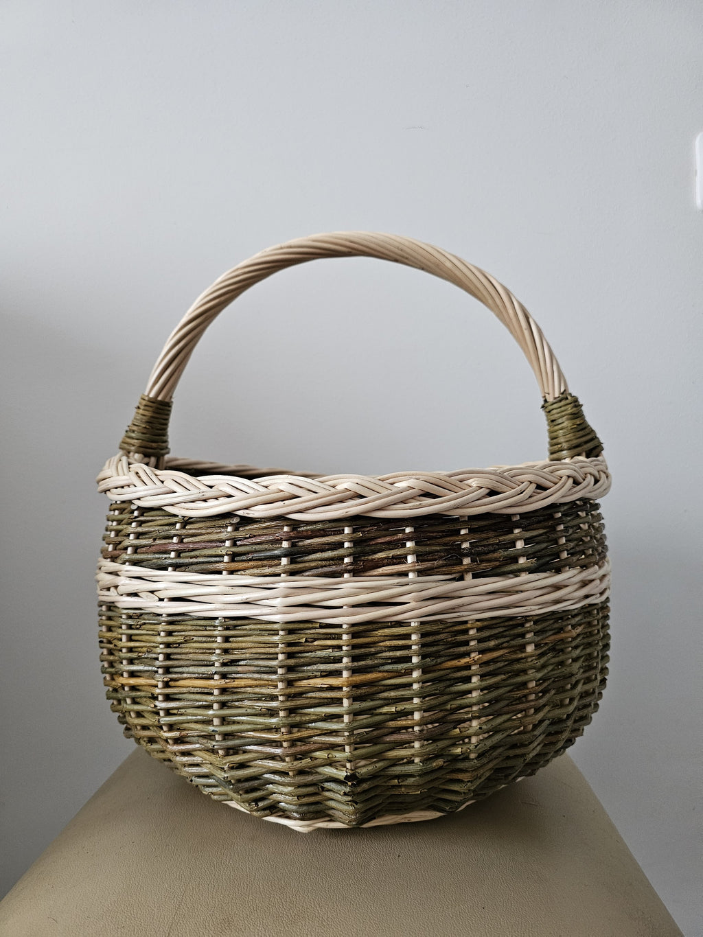 Round willow egg basket with a braided rim, arched handle and a woven band around the body, photographed against a plain grey wall