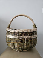 Round willow egg basket with a braided rim, arched handle and a woven band around the body, photographed against a plain grey wall